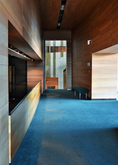 A modern interior hallway featuring wooden paneled walls and a blue carpeted floor. The lighting is soft and ambient, with ceiling fixtures providing additional illumination. A simple bench is situated along one of the walls. The hallway leads to a distant area with more wooden and light-colored wall segments.
