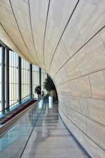 A modern architectural interior with a sweeping, curved wooden wall and tall vertical windows allowing natural light to flood the space. A red-carpeted escalator runs alongside the wall, surrounded by glass railings. Potted plants add greenery to the setting.