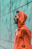 Close-up of a model wearing an international orange unytd hoodie against a black graffiti wall.