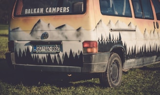 A vintage camper van with a distinctive mountain landscape painted on its exterior. The artwork includes snowy peaks blending into a forest pattern, set against a warm, sunset-colored sky. The van is parked on a grassy field.
