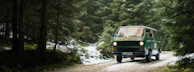 A rugged 4x4 campervan driving along a mountain trail surrounded by deep green forests and towering peaks.