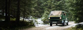 A rugged 4x4 campervan driving along a mountain trail surrounded by deep green forests and towering peaks.