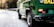 A green van with tropical palm tree graphics on its side is parked on an empty road. The rear of the vehicle shows a yellow print with a logo, and there is a clear view of the taillights. The background features a blurred landscape with trees, suggesting a scenic or natural location.