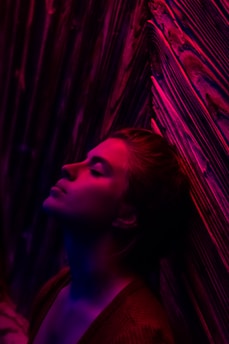 A person is leaning against a wall with eyes closed, illuminated by vivid pink and blue lighting. The wall has a textured appearance with flowing patterns, creating a dreamy and introspective ambiance.