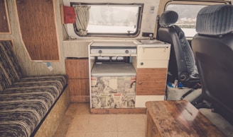 The interior of a camper van featuring a compact kitchen space with a sink and stove. The van has retro-style cabinets and a counter decorated with vintage comic book prints. There is a striped cushioned bench on the left, and the front seats have patterned fabric covers. A small wooden table and a window with a curtain complete the cozy setup.
