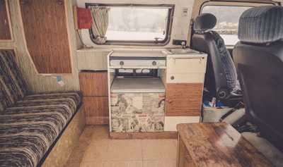 The interior of a camper van featuring a compact kitchen space with a sink and stove. The van has retro-style cabinets and a counter decorated with vintage comic book prints. There is a striped cushioned bench on the left, and the front seats have patterned fabric covers. A small wooden table and a window with a curtain complete the cozy setup.