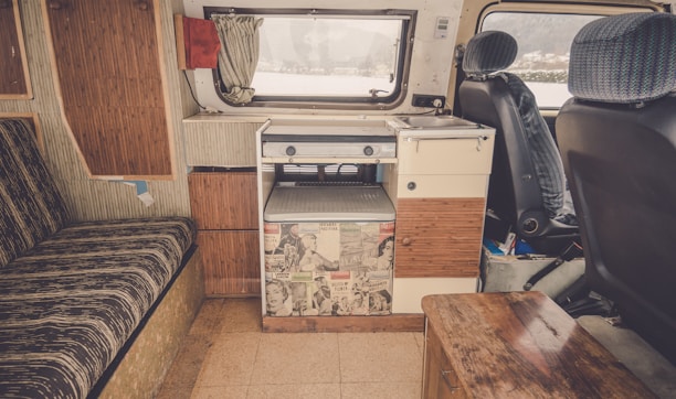 The interior of a camper van featuring a compact kitchen space with a sink and stove. The van has retro-style cabinets and a counter decorated with vintage comic book prints. There is a striped cushioned bench on the left, and the front seats have patterned fabric covers. A small wooden table and a window with a curtain complete the cozy setup.