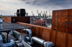 A rooftop scene featuring industrial equipment, with large metal pipes and vents visible in the foreground. The background shows a cityscape with several large cranes towering above warehouses and buildings, indicating an industrial or shipyard area. The sky is partly cloudy, casting a soft light over the entire scene.