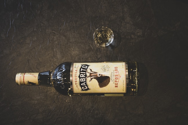 A bottle of Cabrito Reposado tequila lies horizontally on a dark textured surface. There is a small glass filled with a clear liquid positioned near the bottle.