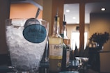 A glass container filled with ice and a metal scoop sits next to several bottles of flavored tequila with pour spouts. The background shows a warm interior setting with soft lighting.