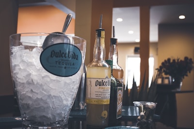 A glass container filled with ice and a metal scoop sits next to several bottles of flavored tequila with pour spouts. The background shows a warm interior setting with soft lighting.