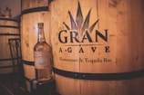 A bottle of Casamigos tequila is placed on a small box in front of large wooden barrels with the label 'El Gran Agave Restaurant & Tequila Bar' printed on them. The setting appears to be inside a distillery or themed bar featuring rustic wooden textures.