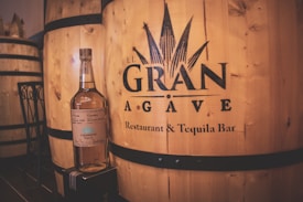 A bottle of Casamigos tequila is placed on a small box in front of large wooden barrels with the label 'El Gran Agave Restaurant & Tequila Bar' printed on them. The setting appears to be inside a distillery or themed bar featuring rustic wooden textures.