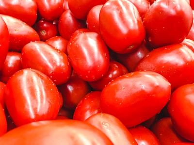 A large pile of ripe, red Roma tomatoes fills the frame, showcasing their smooth, shiny, and slightly oval textures. The tomatoes are tightly packed together, creating a vibrant and fresh appearance.