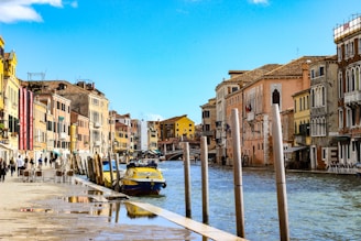 boat near sea and people walking on pathway beside buildings under blue and white skies