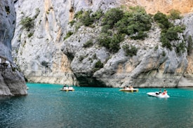 Pedal boats navigate the turquoise waters of a narrow canyon flanked by steep limestone cliffs. The cliffs are dotted with patches of greenery, adding contrast to the rocky surfaces.
