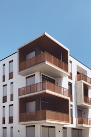 A modern apartment building featuring multiple stories with prominent geometric designs. The structure showcases large balconies with wooden or metal railings, and the exterior is painted in a combination of white and earthy tones. The sky is clear and blue, enhancing the minimalist and contemporary architectural style.