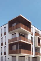 A modern apartment building featuring multiple stories with prominent geometric designs. The structure showcases large balconies with wooden or metal railings, and the exterior is painted in a combination of white and earthy tones. The sky is clear and blue, enhancing the minimalist and contemporary architectural style.