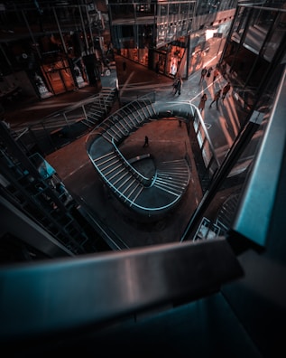 A modern architectural interior with a spiraling staircase and glass-walled structures. People are visible walking near the structures, creating dynamic shadows on the floor. The space has an urban and futuristic aesthetic with reflections on the glass surfaces.