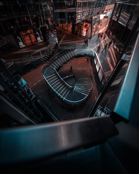 A modern architectural interior with a spiraling staircase and glass-walled structures. People are visible walking near the structures, creating dynamic shadows on the floor. The space has an urban and futuristic aesthetic with reflections on the glass surfaces.