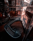 A modern interior space featuring a spiral staircase, with reflections and shadows on glass surfaces. The ambient lighting creates a warm, inviting atmosphere. Shoppers can be seen walking around, with stores lining the surroundings.