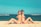 Two women in swimsuits are sitting back-to-back on a sandy beach with the ocean in the background. The woman on the left is wearing a white bikini while the woman on the right is in a red bikini and sunglasses. The sky is clear with a few clouds, and the overall atmosphere is sunny and relaxed.