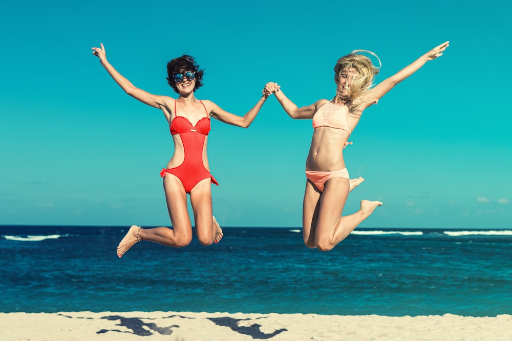 two woman in swimsuit jumping on seashore during daytime