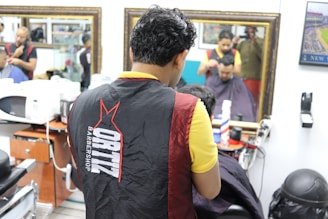 Multiple barbers are cutting hair inside a barbershop with mirrors on the walls. The focus is on a barber in a black and red apron working on a customer. There are hair styling tools on counters. The environment is bright and busy, with reflections of people in the mirrors and framed pictures on the walls.