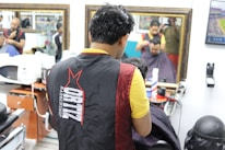 Multiple barbers are cutting hair inside a barbershop with mirrors on the walls. The focus is on a barber in a black and red apron working on a customer. There are hair styling tools on counters. The environment is bright and busy, with reflections of people in the mirrors and framed pictures on the walls.