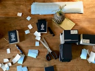 A wooden table is cluttered with stationery items, including several stacks of small notebooks that have 'I WILL KEEP MY WORD' printed on them. There are scattered blue and white labels or stickers, a pair of black-handled scissors, a pen, and a decorative vase with dried grass.