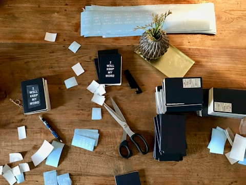 A wooden table is cluttered with stationery items, including several stacks of small notebooks that have 'I WILL KEEP MY WORD' printed on them. There are scattered blue and white labels or stickers, a pair of black-handled scissors, a pen, and a decorative vase with dried grass.