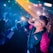 A performer on stage is actively engaging with the audience, holding a microphone and wearing sunglasses. The performer is dressed in a colorful jacket with pink and blue hues. There are bright lights and the audience is capturing the moment with their phones, adding to the vibrant atmosphere.