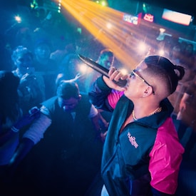 A performer on stage is actively engaging with the audience, holding a microphone and wearing sunglasses. The performer is dressed in a colorful jacket with pink and blue hues. There are bright lights and the audience is capturing the moment with their phones, adding to the vibrant atmosphere.