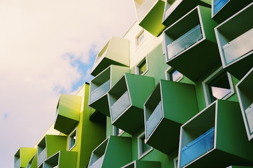 A modern building with protruding green cubic balconies against a partly cloudy sky. The architectural design emphasizes geometric shapes and a vibrant green color palette.