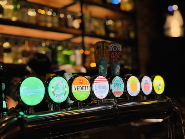 A bar setting features multiple beer taps with illuminated logos, including brands such as Carlsberg, Vedett, and Union Jack IPA. The background is dimly lit with shelves of bottles and glasses, creating a cozy and inviting atmosphere.