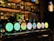 A bar setting features multiple beer taps with illuminated logos, including brands such as Carlsberg, Vedett, and Union Jack IPA. The background is dimly lit with shelves of bottles and glasses, creating a cozy and inviting atmosphere.