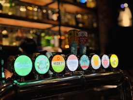 A bar setting features multiple beer taps with illuminated logos, including brands such as Carlsberg, Vedett, and Union Jack IPA. The background is dimly lit with shelves of bottles and glasses, creating a cozy and inviting atmosphere.