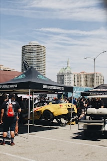 A vibrant outdoor car event features a yellow sports car under a black canopy with the signage 'DEEP LIFESTYLES SUPPLY CO.' People gather around the car, suggesting an exhibition or a car show. In the background, tall buildings rise under a partly cloudy sky. Additional tents and setups are visible, indicating a bustling atmosphere.