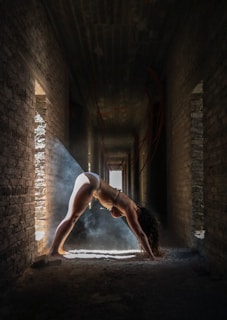 A person is performing a downward-facing yoga pose in the middle of a narrow, dimly lit corridor. A ray of sunlight streams through a side window, casting a dramatic beam that highlights the figure amidst a backdrop of textured brick walls. The ambiance suggests a blend of serenity and tension, with the light smoke or dust particles enhancing the ethereal quality of the scene.