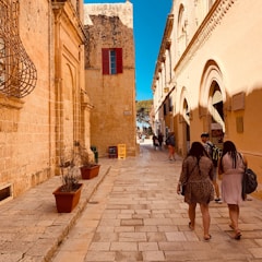 A narrow cobblestone street lined with historical stone buildings. There are people walking along the street, and one building displays an ATM sign. Potted plants are placed along the ledge of one building with a rustic latticed wooden structure attached to it.
