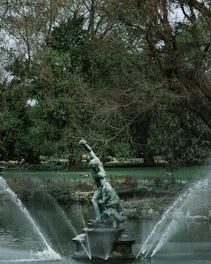 A classical statue positioned centrally in a serene garden setting with water fountains. The sculpture depicts a muscular figure in a dynamic pose on a pedestal, surrounded by a pond. Trees with dense foliage frame the scene, casting an aura of tranquility.