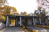 The entrance of a university campus features a structure with a sign that reads 'Ankara Üniversitesi Cebeci Yerleşkesi'. Tall trees with yellow and green foliage are visible on either side of the road leading up to the building. Several people are walking in and out of the entrance, suggesting activity and life on campus. The sunlight filters through the trees, casting a warm glow over the scene.