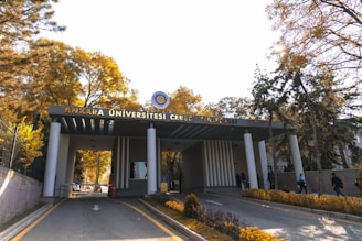 The entrance of a university campus features a structure with a sign that reads 'Ankara Üniversitesi Cebeci Yerleşkesi'. Tall trees with yellow and green foliage are visible on either side of the road leading up to the building. Several people are walking in and out of the entrance, suggesting activity and life on campus. The sunlight filters through the trees, casting a warm glow over the scene.