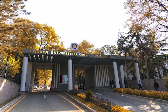 The entrance of a university campus features a structure with a sign that reads 'Ankara Üniversitesi Cebeci Yerleşkesi'. Tall trees with yellow and green foliage are visible on either side of the road leading up to the building. Several people are walking in and out of the entrance, suggesting activity and life on campus. The sunlight filters through the trees, casting a warm glow over the scene.