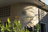 A group of people stand in front of a building with the inscription 'Ankara Üniversitesi Dil ve Tarih-Coğrafya Fakültesi' on the wall. The building facade is made of stone tiles, and there are some green plants in the foreground casting shadows.