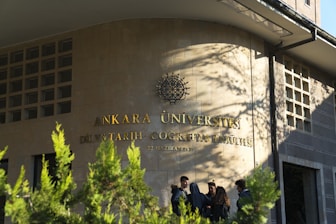 A group of people stand in front of a building with the inscription 'Ankara Üniversitesi Dil ve Tarih-Coğrafya Fakültesi' on the wall. The building facade is made of stone tiles, and there are some green plants in the foreground casting shadows.