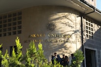 A group of people stand in front of a building with the inscription 'Ankara Üniversitesi Dil ve Tarih-Coğrafya Fakültesi' on the wall. The building facade is made of stone tiles, and there are some green plants in the foreground casting shadows.