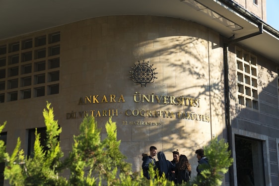 A group of people stand in front of a building with the inscription 'Ankara Üniversitesi Dil ve Tarih-Coğrafya Fakültesi' on the wall. The building facade is made of stone tiles, and there are some green plants in the foreground casting shadows.