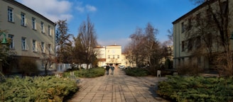 A pathway stretches between two buildings under a blue sky with some clouds. There are bushes and trees on both sides of the pathway. In the distance, two people are walking towards a building with a large clock above its entrance.