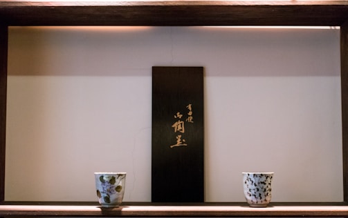 A wooden shelf features two ceramic cups with decorative patterns placed symmetrically on either side of a vertical wooden plaque with Japanese characters. The setting is minimalistic and arranged against a light-colored wall.